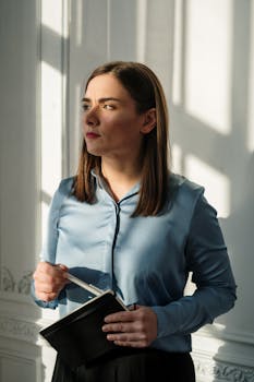 A contemplative woman stands indoors, holding a notebook, bathed in warm sunlight.