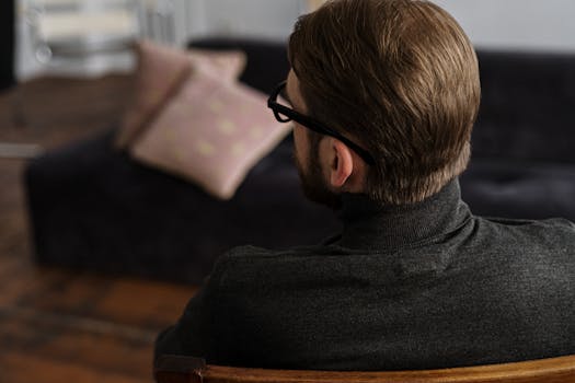 A man with glasses sits contemplatively indoors, suggesting a reflective or therapeutic moment.