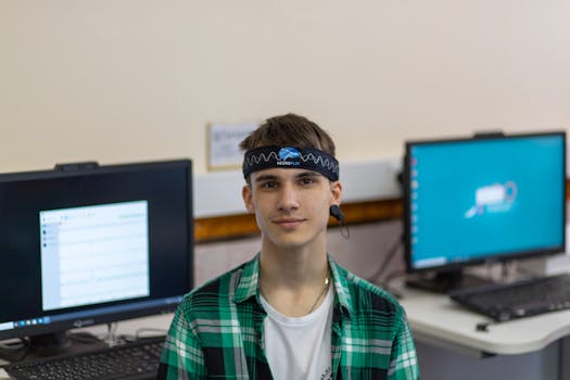 Portrait of a young man in a plaid shirt wearing a Neuroplay headband with computers in the background.