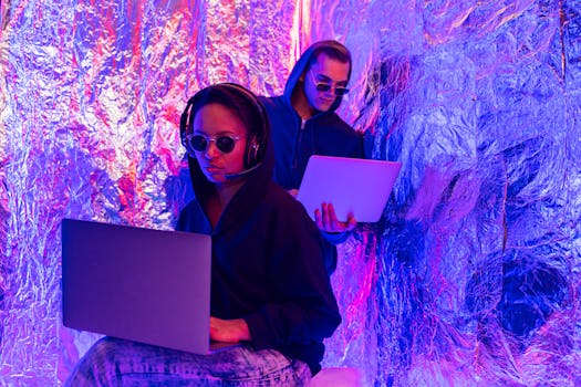 Two individuals wearing hoodies working on laptops in a neon-lit, futuristic room.