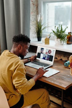 African American man using tablet for video call at home desk with laptop.